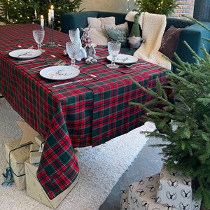 May include: A festive Christmas table setting with a red and green plaid tablecloth. The table is set with plates, silverware, and crystal glasses. Decorative elements include candles, gnomes, and small Christmas trees. Presents are wrapped and placed under the table.