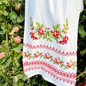 K&ouml;nnte beinhalten: Wei&szlig;es Leinenhandtuch mit gestickten roten Beeren, gr&uuml;nen Bl&auml;ttern und braunen Tannenzapfen. Der untere Rand hat ein rotes geometrisches Muster. Das Handtuch h&auml;ngt an einem Ast mit &Auml;pfeln.