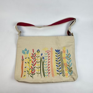Puede incluir: Una bolsa de lona beige con una correa de hombro roja y un dise&ntilde;o floral bordado en tonos de azul, amarillo, verde y rojo.