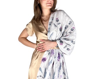 May include: A white linen scarf with a floral design in shades of purple and blue. The scarf is draped around a person's shoulders and tied at the waist.