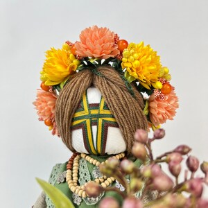 Puede incluir: Mu&ntilde;eca hecha a mano con una corona floral. La mu&ntilde;eca tiene cabello de hilo marr&oacute;n y una cara blanca con detalles bordados. Viste un vestido verde, un chal beige y un collar de cuentas. La corona presenta flores naranjas y amarillas.