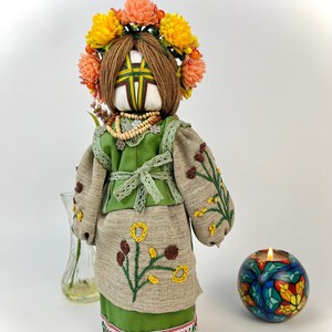 Puede incluir: Mu&ntilde;eca hecha a mano con un vestido bordado verde y beige, una corona floral y un collar de cuentas. La mu&ntilde;eca tiene cabello de hilo marr&oacute;n y est&aacute; adornada con bordados florales amarillos y marrones. Un candelabro y un jarr&oacute;n est&aacute;n en el fondo.