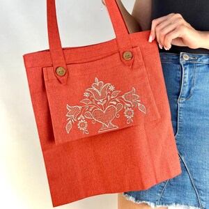 Op de afbeelding: Een rode tote bag met een wit geborduurd bloemenontwerp en twee houten knopen. De tas heeft een groot vak aan de voorkant.