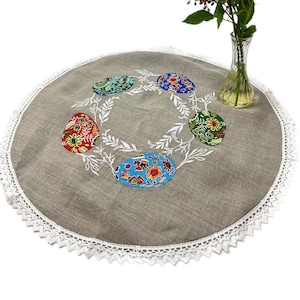 May include: A round, beige linen tablecloth with a white lace trim. The tablecloth features an embroidered design of four colourful Easter eggs surrounded by white leaves.
