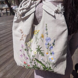 Op de afbeelding: Een beige canvas draagtas met een wit touw handvat en geborduurde wilde bloemen in tinten van blauw, geel, groen en roze.