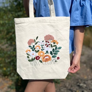 May include: A white canvas tote bag with a floral embroidery design. The embroidery features a variety of flowers in shades of pink, red, orange, yellow, and green.