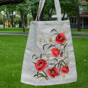 Op de afbeelding: Een linnen tas met geborduurde rode klaprozen en witte madeliefjes. De tas heeft een natuurlijke linnen kleur en hangt aan een tak.