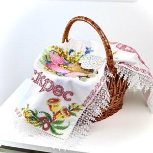May include: A wicker basket with a handle, containing a white embroidered cloth. The cloth is decorated with colorful floral and religious embroidery, and has a white lace trim. The basket sits on a white surface.