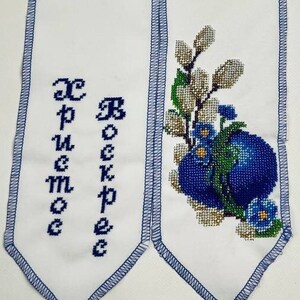 Könnte beinhalten: Weißes Stoffband mit blauen bestickten Kanten. Das Band hat zwei Enden mit besticktem Text in kyrillischer Schrift: "С Рождеством" und einem blauen und weißen bestickten Blumendesign mit einem blauen Ei.