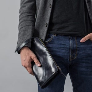 May include: A person wearing a gray blazer, black t-shirt, blue jeans, and a silver watch. They are holding a black leather clutch.