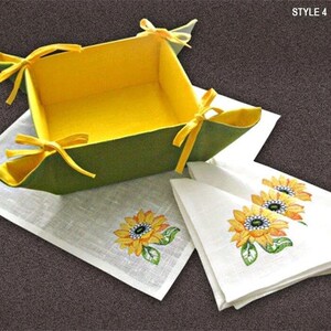 May include: A green and yellow felt bread basket with yellow ribbon ties. The basket is sitting on a white linen placemat with embroidered sunflowers. Three white linen napkins with embroidered sunflowers are folded next to the basket. The text "STYLE 4" is in the upper right corner of the image.