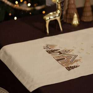 Op de afbeelding: Een beige tafelkleed met een geborduurd ontwerp van peperkoekhuisjes en sneeuw. De tafelkleed ligt op een bruine tafel met een gouden rendierfiguur en een kom met dennenappels.