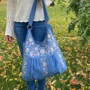 Peut inclure: Un sac cabas bleu avec des motifs floraux brod&eacute;s blancs. Le sac a un haut large et ouvert et deux longues anses.