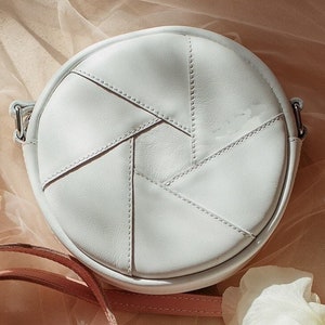 May include: A white round leather crossbody bag with a geometric design. The bag has a single strap and is made of stitched leather panels.