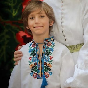 May include: A woman and a young boy wearing traditional Ukrainian embroidered shirts, or vyshyvankas. The woman is wearing a white linen shirt with a green belt and gold earrings. The boy is wearing a white shirt with a colorful floral embroidery pattern and a blue tassel. They are standing in front of a floral patterned background.