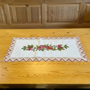 May include: A white crocheted table runner with a red berry and green leaf embroidery design on a wooden table.