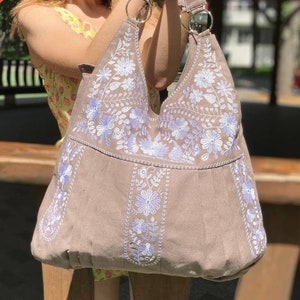K&ouml;nnte beinhalten: Eine taupefarbene Umh&auml;ngetasche mit wei&szlig;en und hellvioletten Blumenstickereien. Die Tasche hat einen breiten Riemen und Metallringe. Die Tasche wird von einer Person gehalten.