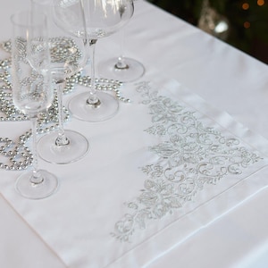 May include: A white tablecloth with a decorative embroidered runner and several wine glasses. The runner features an ornate silver floral design. Silver beaded garland is draped across the table, creating a festive holiday setting.