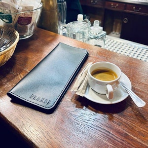 K&ouml;nnte beinhalten: Eine schwarze Lederkarte mit dem gepr&auml;gten Wort "Flaxsi" auf der Vorderseite, eine wei&szlig;e Tasse Kaffee auf einem wei&szlig;en Untersetzer und ein P&auml;ckchen Zuckerst&auml;bchen auf einem Holztisch.