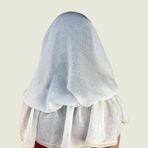 May include: A white, textured fabric head covering and cape. The cape has a ruffled bottom edge with a cream-colored lace trim. The head covering drapes over the head and shoulders.