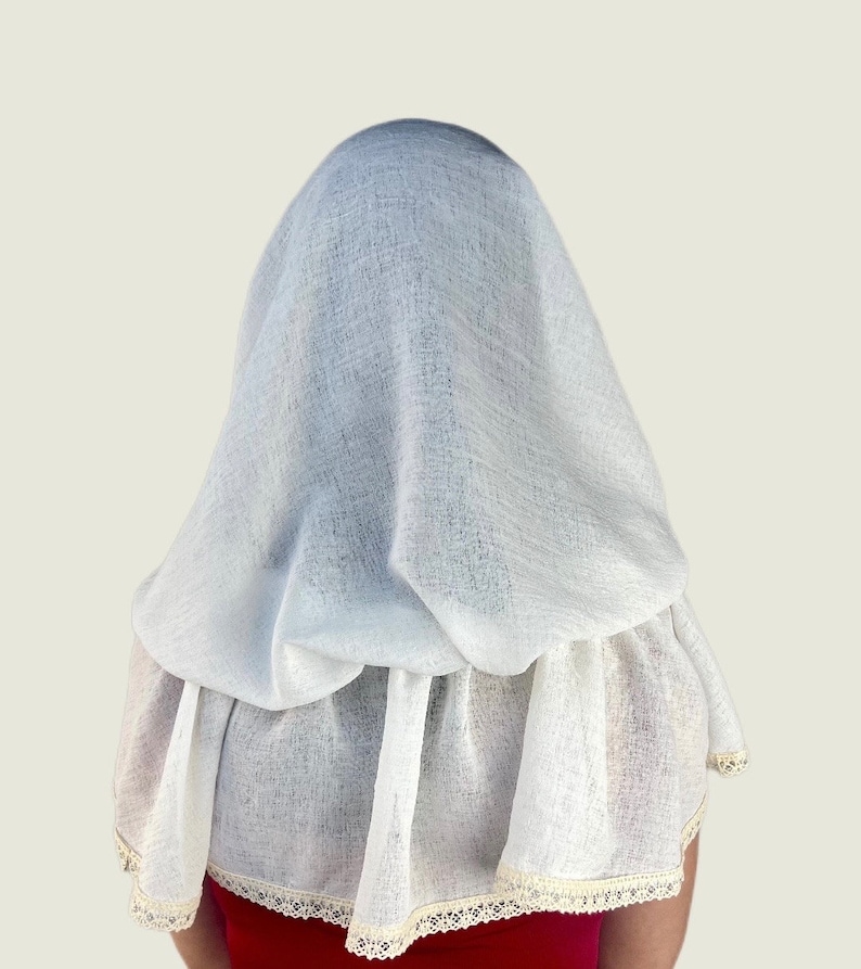 May include: A white, textured fabric head covering and cape. The cape has a ruffled bottom edge with a cream-colored lace trim. The head covering drapes over the head and shoulders.