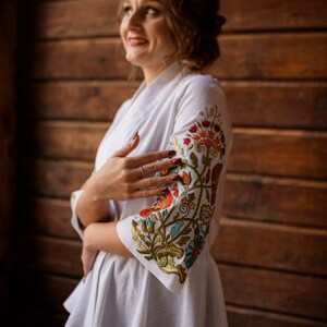 Op de afbeelding: Een wit geborduurd blouse met een bloemenpatroon in tinten rood, oranje, groen, blauw en goud. De blouse heeft een gerimpelde zoom en driekwart mouwen.
