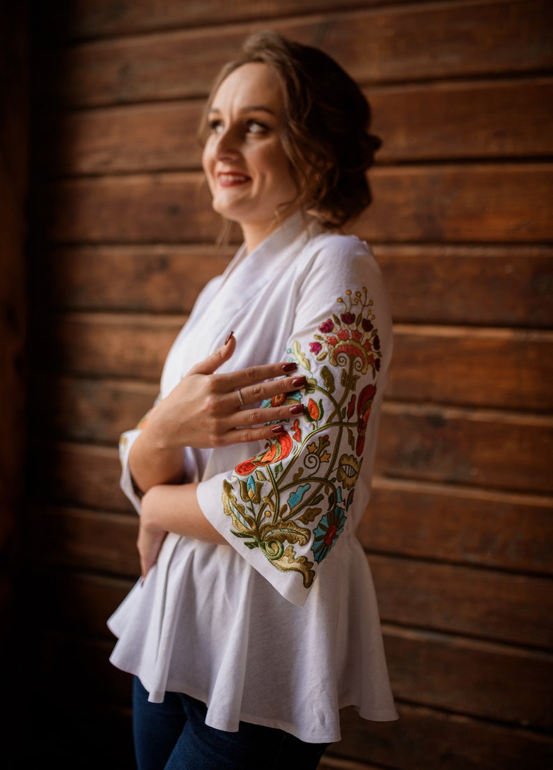 Op de afbeelding: Een wit geborduurd blouse met een bloemenpatroon in tinten rood, oranje, groen, blauw en goud. De blouse heeft een gerimpelde zoom en driekwart mouwen.
