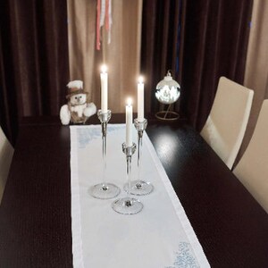 May include: A festive dining table setting with a white runner featuring a blue snowflake design. Three crystal candle holders with lit candles are centered on the runner. A Christmas wreath and ornament add to the holiday theme.