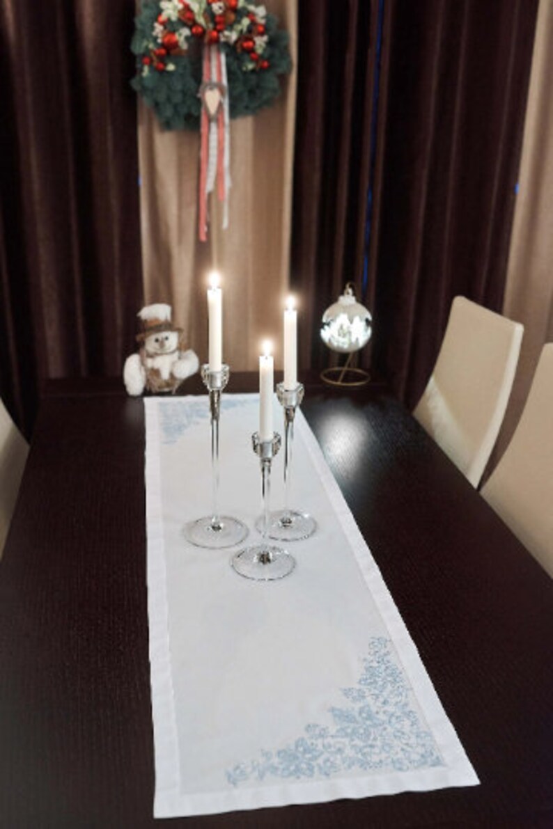 May include: A festive dining table setting with a white runner featuring a blue snowflake design. Three crystal candle holders with lit candles are centered on the runner. A Christmas wreath and ornament add to the holiday theme.