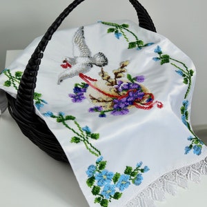 May include: White embroidered cloth featuring a dove, flowers, and a basket design. The cloth has a lace trim and is in a black wicker basket.