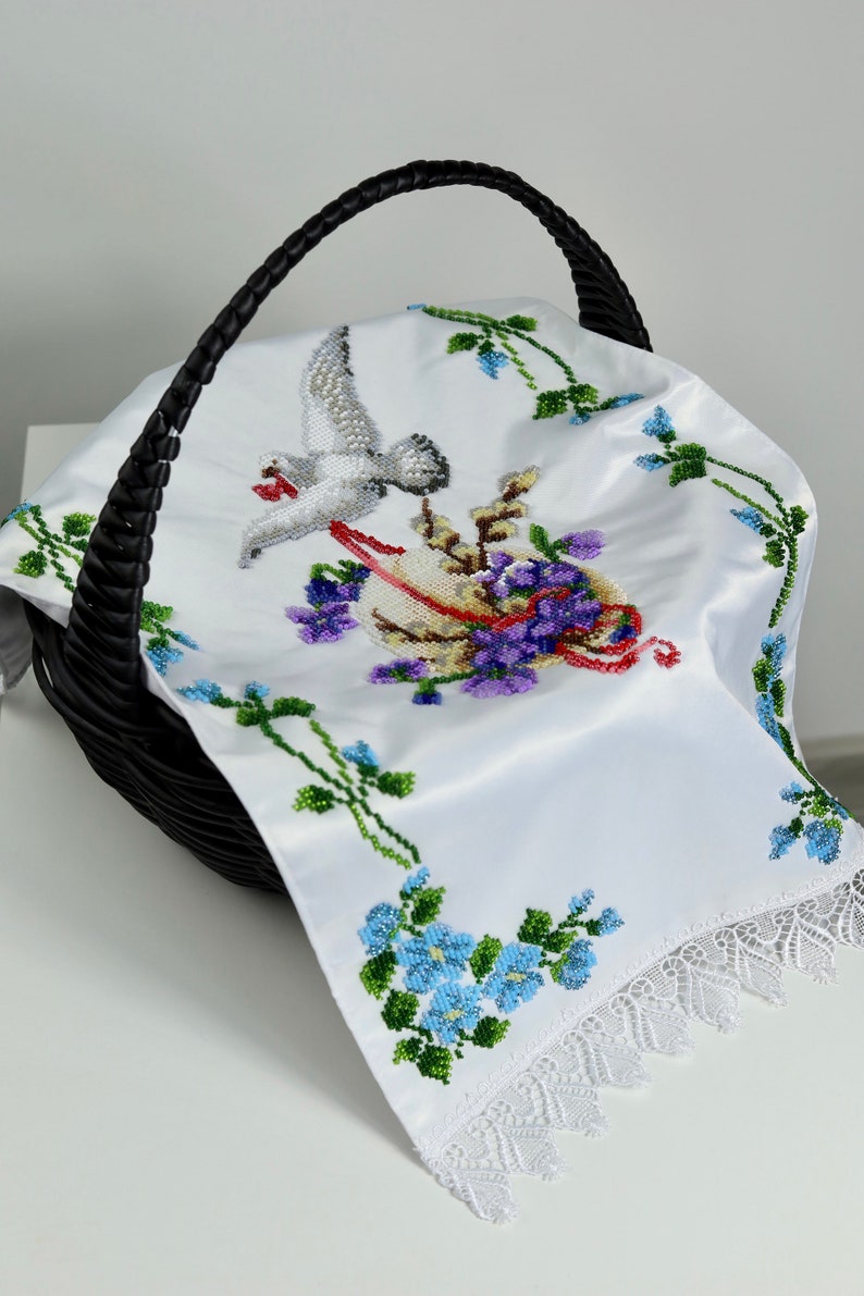 May include: White embroidered cloth featuring a dove, flowers, and a basket design. The cloth has a lace trim and is in a black wicker basket.