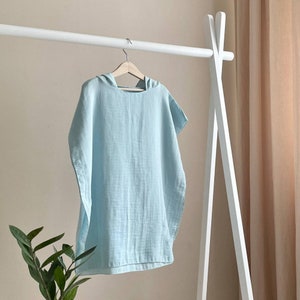 Puede incluir: Una toalla con capucha de color azul claro colgada de un estante de madera blanco. La toalla est&aacute; hecha de un tejido suave y ligero.