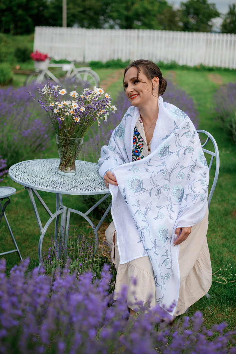 Puede incluir: Una mujer con un chal blanco con un dise&ntilde;o floral azul, sentada en una peque&ntilde;a mesa de metal. La escena est&aacute; ambientada en un campo de lavanda, con un jarr&oacute;n de flores en la mesa y una bicicleta blanca al fondo.