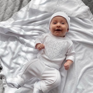 May include: A white baby outfit with a cross embroidered on the chest. The outfit includes a long-sleeved shirt, pants, and a hat. The baby is lying on a white blanket with a lace trim.