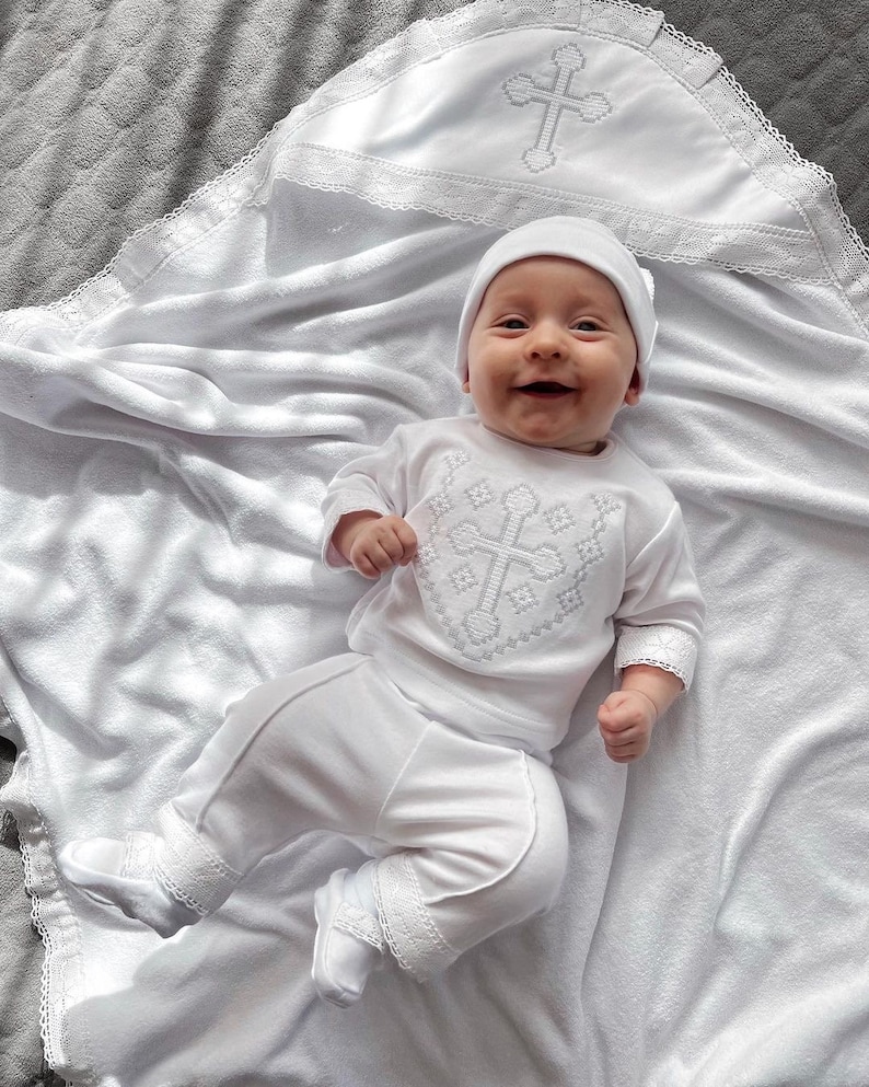 May include: A white baby outfit with a cross embroidered on the chest. The outfit includes a long-sleeved shirt, pants, and a hat. The baby is lying on a white blanket with a lace trim.