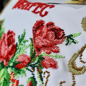K&ouml;nnte beinhalten: Nahaufnahme eines wei&szlig;en Stoffes mit aufwendiger Perlenstickerei. Das Design zeigt gro&szlig;e rote Rosen, Knospen und gr&uuml;ne Bl&auml;tter. Die Perlen sind in Rot-, Rosa-, Gr&uuml;n- und Goldt&ouml;nen gehalten und bilden ein strukturiertes Blumenmuster. Das Wort "MARY" ist ebenfalls rot aufgestickt.