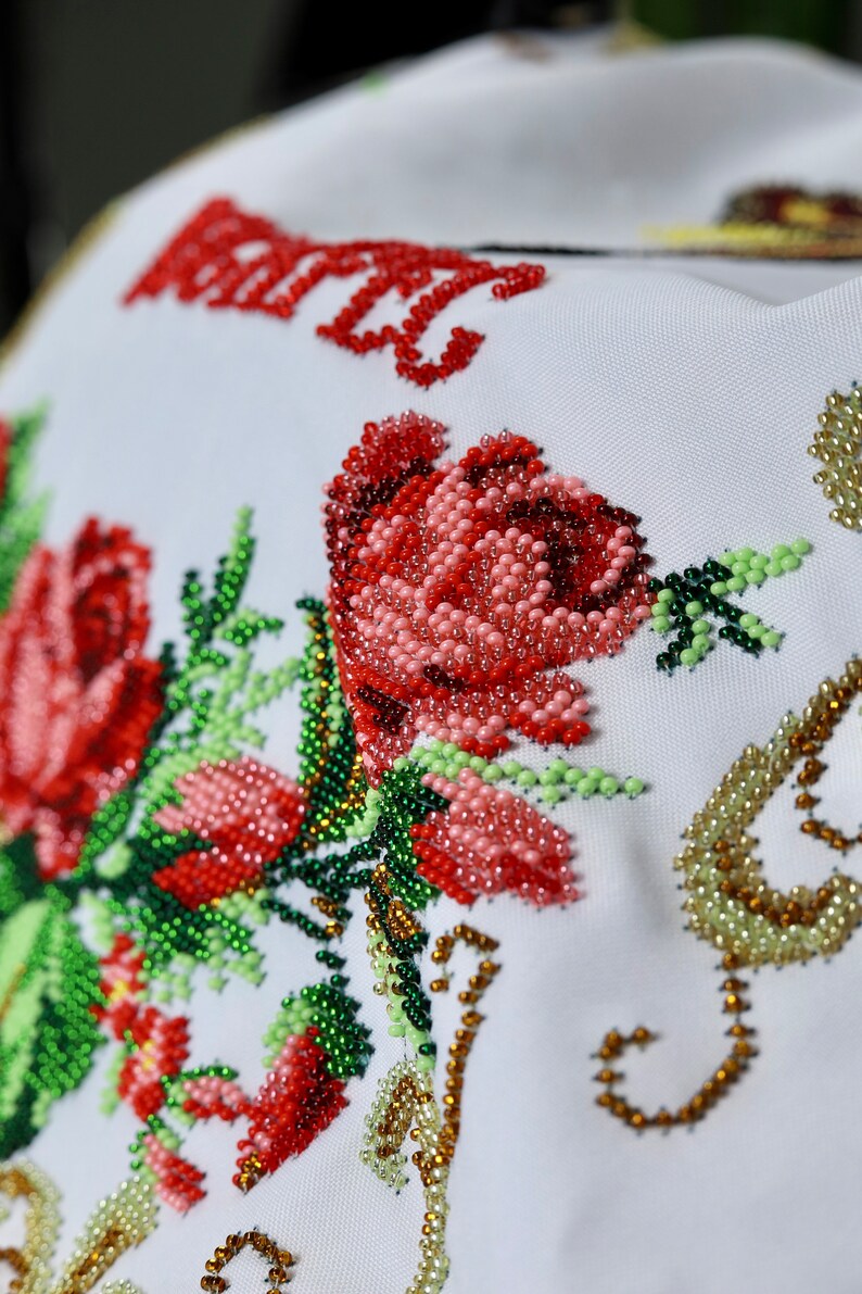 K&ouml;nnte beinhalten: Nahaufnahme eines wei&szlig;en Stoffes mit aufwendiger Perlenstickerei. Das Design zeigt gro&szlig;e rote Rosen, Knospen und gr&uuml;ne Bl&auml;tter. Die Perlen sind in Rot-, Rosa-, Gr&uuml;n- und Goldt&ouml;nen gehalten und bilden ein strukturiertes Blumenmuster. Das Wort "MARY" ist ebenfalls rot aufgestickt.