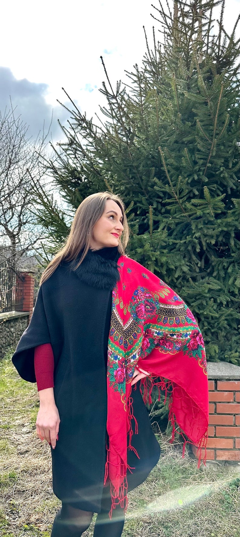 May include: A woman wearing a black coat with a fur collar and a red shawl with floral embroidery. The shawl has fringe trim and is draped over her shoulder.