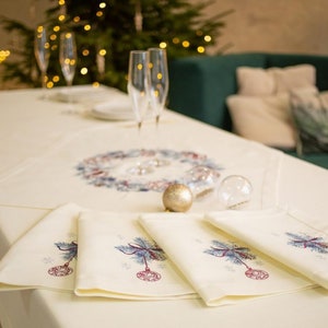 Puede incluir: Una mesa festiva con un mantel y servilletas color crema. Las servilletas presentan bordados de adornos navide&ntilde;os. Se observa un centro de mesa con una corona y un adorno dorado, junto a copas de champ&aacute;n y un &aacute;rbol de Navidad al fondo.