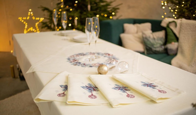 Puede incluir: Una mesa festiva con un mantel y servilletas color crema. Las servilletas presentan bordados de adornos navide&ntilde;os. Se observa un centro de mesa con una corona y un adorno dorado, junto a copas de champ&aacute;n y un &aacute;rbol de Navidad al fondo.