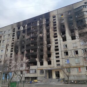 K&ouml;nnte beinhalten: Au&szlig;enansicht eines mehrst&ouml;ckigen Wohngeb&auml;udes mit erheblichen Brandsch&auml;den. Die Fassade ist grau, mit ausgedehnten schwarzen Verkohlungen auf mehreren Etagen, was auf einen Brand hindeutet. Fenster sind zerbrochen oder fehlen.