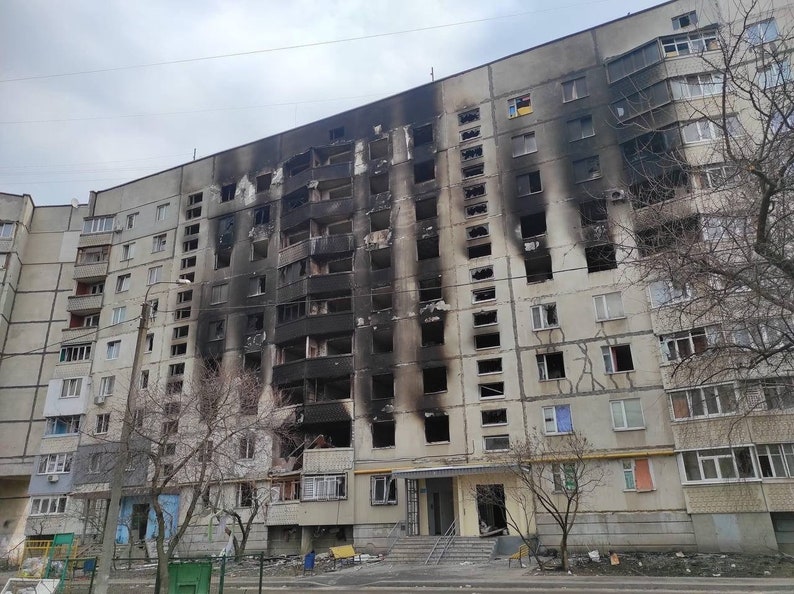 K&ouml;nnte beinhalten: Au&szlig;enansicht eines mehrst&ouml;ckigen Wohngeb&auml;udes mit erheblichen Brandsch&auml;den. Die Fassade ist grau, mit ausgedehnten schwarzen Verkohlungen auf mehreren Etagen, was auf einen Brand hindeutet. Fenster sind zerbrochen oder fehlen.
