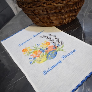 Puede incluir: Toalla de t&eacute; de lino blanco con ribete azul bordado. La toalla presenta un dise&ntilde;o floral con narcisos amarillos, nomeolvides azules y ramas de sauce grises. El texto "Христос Воскрес" y "Воистину Воскрес" est&aacute; bordado en hilo azul.