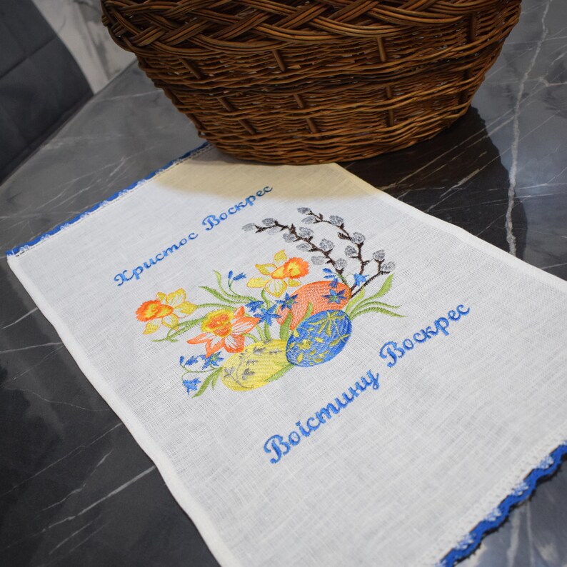 Puede incluir: Toalla de t&eacute; de lino blanco con ribete azul bordado. La toalla presenta un dise&ntilde;o floral con narcisos amarillos, nomeolvides azules y ramas de sauce grises. El texto "Христос Воскрес" y "Воистину Воскрес" est&aacute; bordado en hilo azul.