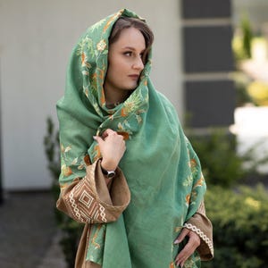 May include: A woman wearing a green shawl with floral embroidery. The shawl is draped over her head and shoulders, and she is looking to the side. She is wearing a brown dress with embroidered details.