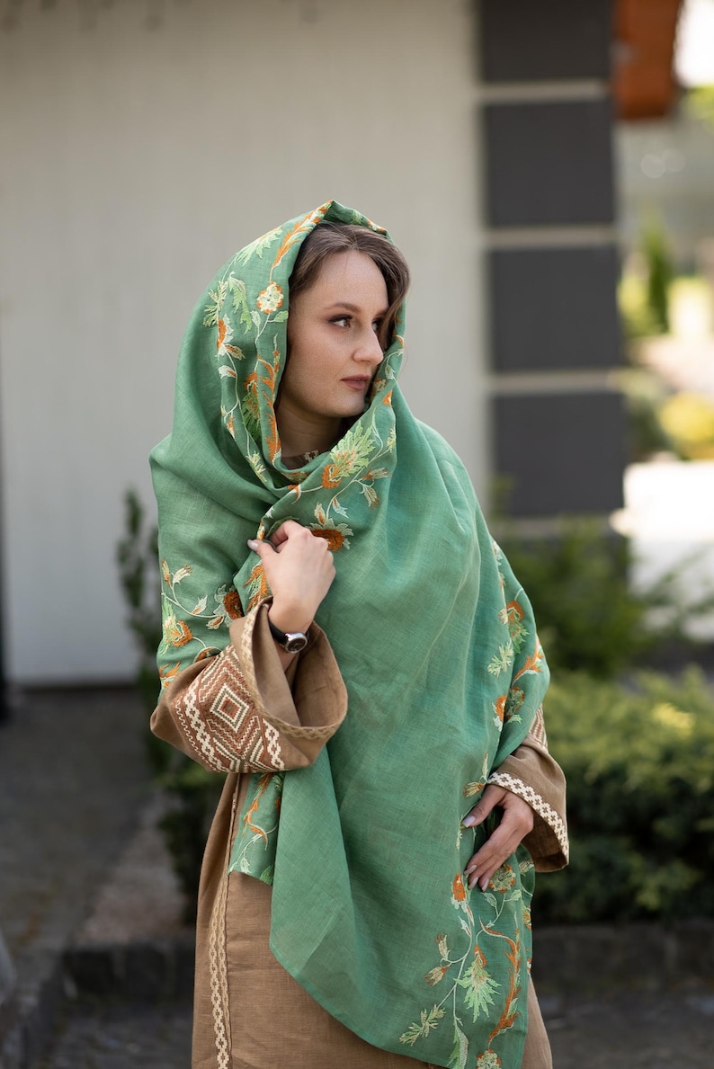 May include: A woman wearing a green shawl with floral embroidery. The shawl is draped over her head and shoulders, and she is looking to the side. She is wearing a brown dress with embroidered details.