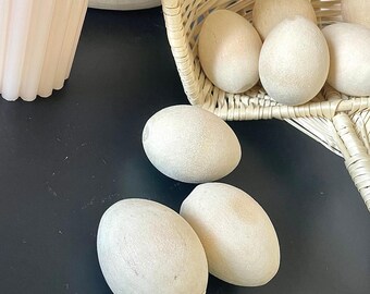 May include: A light-colored woven basket filled with several smooth, matte-finish egg-shaped objects. Three additional egg-shaped objects are positioned outside the basket on a dark surface, displaying a uniform, pale, neutral tone.