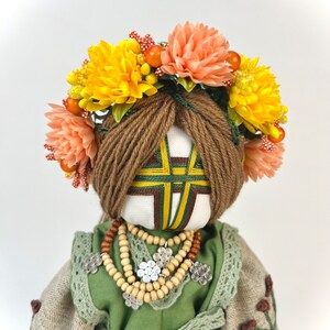 Puede incluir: Mu&ntilde;eca hecha a mano con una corona floral de flores amarillas y melocot&oacute;n. La mu&ntilde;eca tiene cabello de hilo marr&oacute;n, un vestido verde y beige y un collar de cuentas de madera. La cara de la mu&ntilde;eca tiene un dise&ntilde;o geom&eacute;trico.
