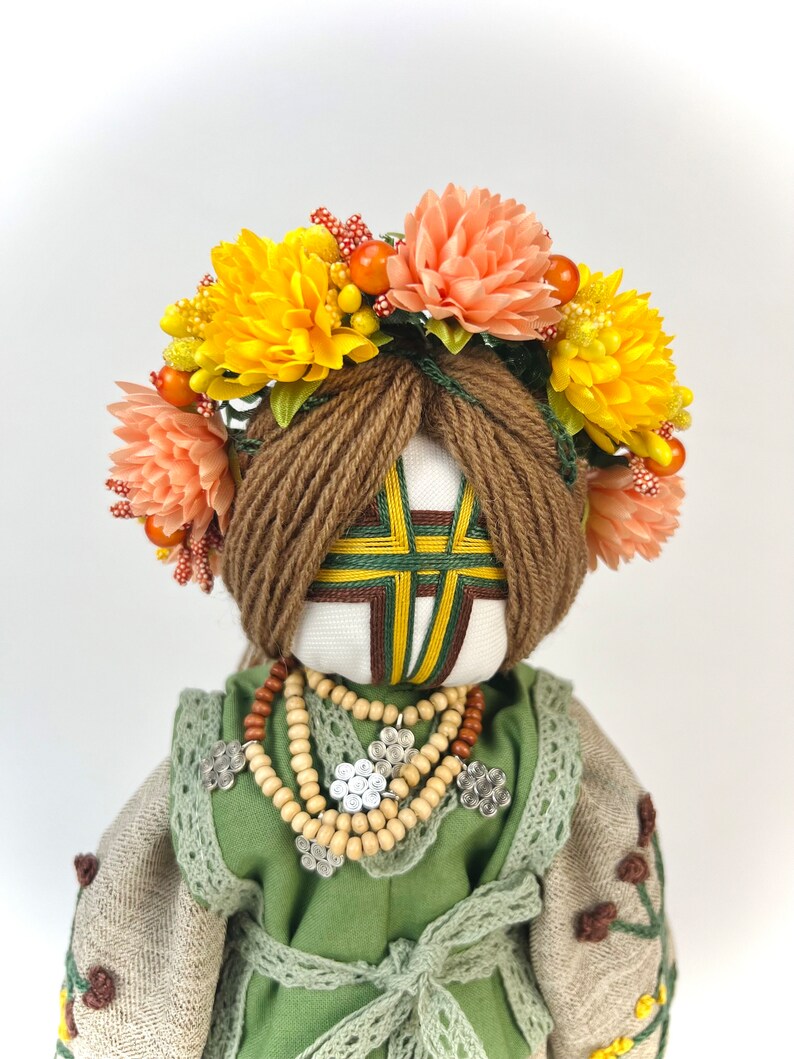 Puede incluir: Mu&ntilde;eca hecha a mano con una corona floral de flores amarillas y melocot&oacute;n. La mu&ntilde;eca tiene cabello de hilo marr&oacute;n, un vestido verde y beige y un collar de cuentas de madera. La cara de la mu&ntilde;eca tiene un dise&ntilde;o geom&eacute;trico.