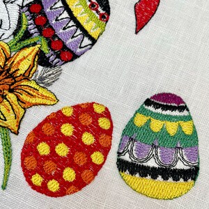 May include: Three embroidered Easter eggs with different patterns. One egg is purple, green, and yellow with a black and white border. Another egg is red and yellow with a polka dot pattern. The third egg is purple, yellow, and green with a zig-zag pattern.